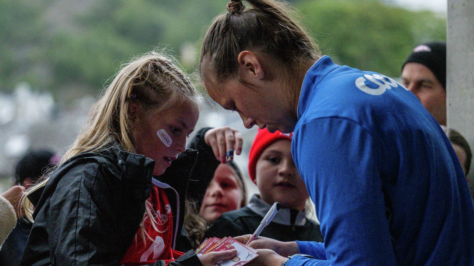 En kvinnelig idrettsutøver i en blå trøye signerer autografer for unge fans på et utendørsarrangement.