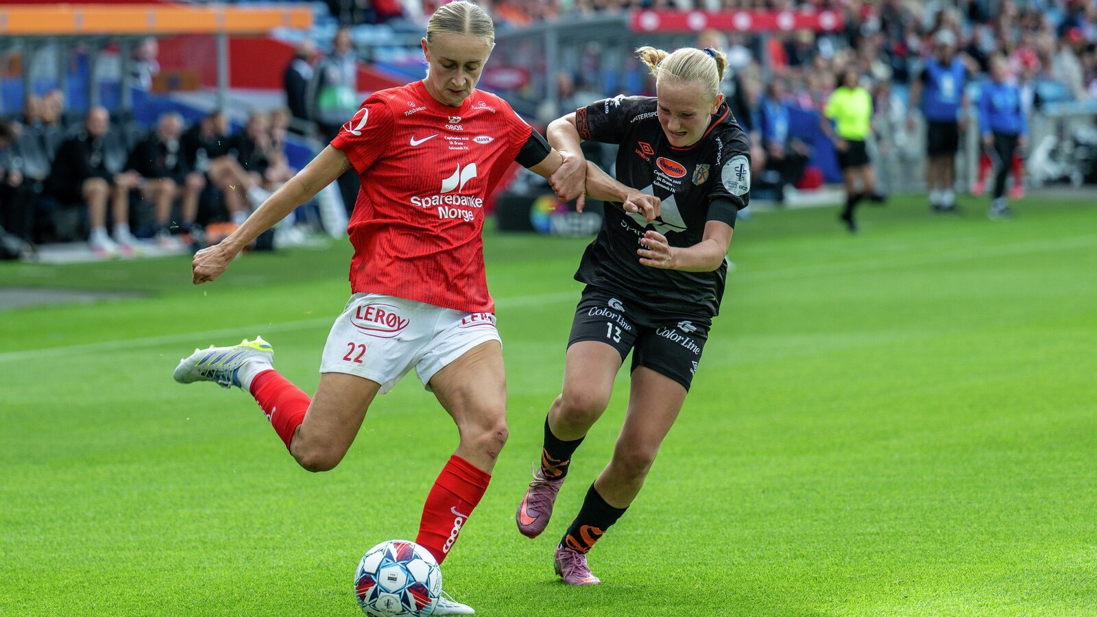 To kvinnelige fotballspillere konkurrerer om ballen under en kamp, iført røde og svarte uniformer.