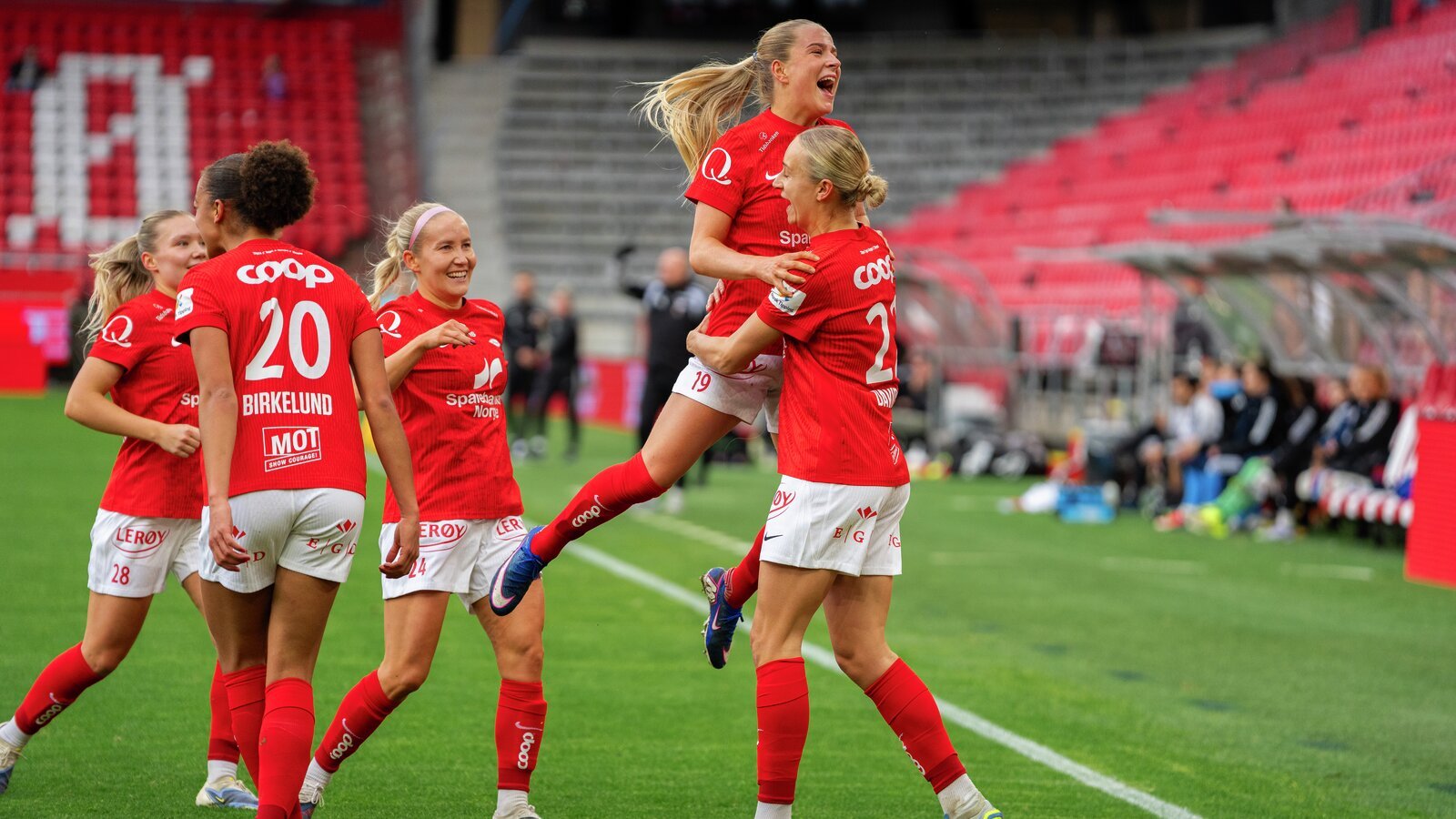 Kvinnelige fotballspillere i røde uniformer feirer et mål på banen, med tomme stadionseter i bakgrunnen.