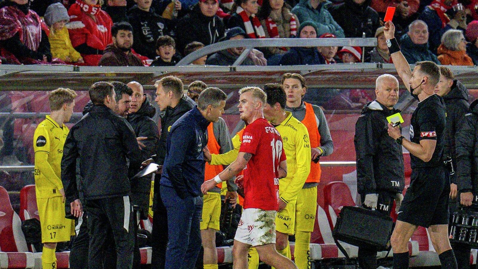 Fotballspillere og dommere samlet seg på banen under en kamp, med røde og gule drakter synlige.