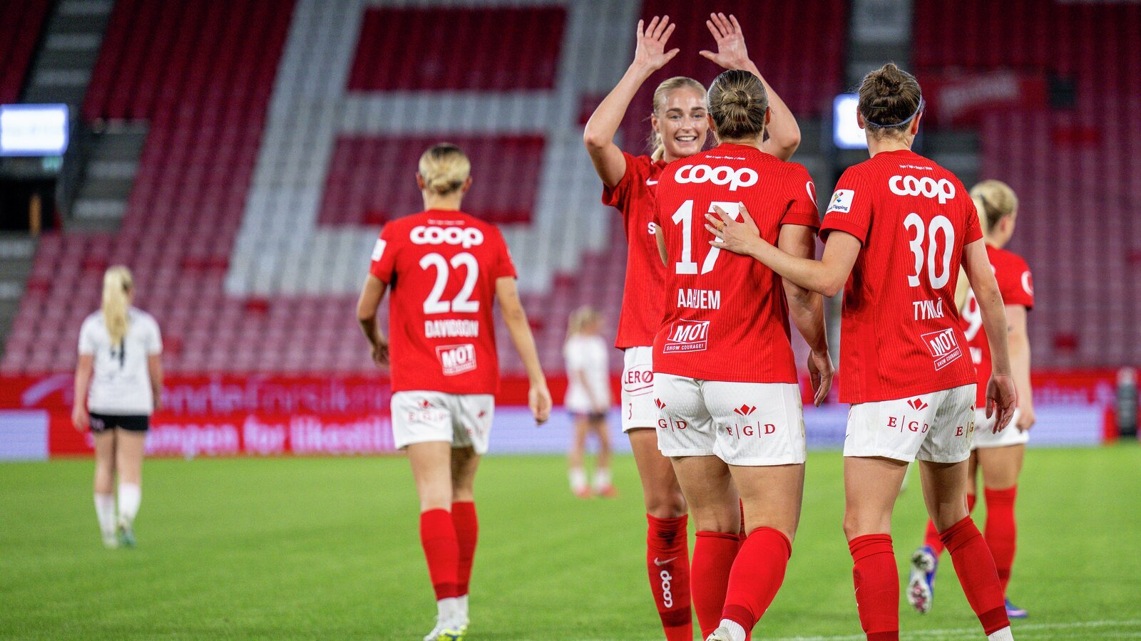 Kvinnefotballspillere i røde COOP-drakter feirer på banen, med numrene 22, 17 og 30 synlige.