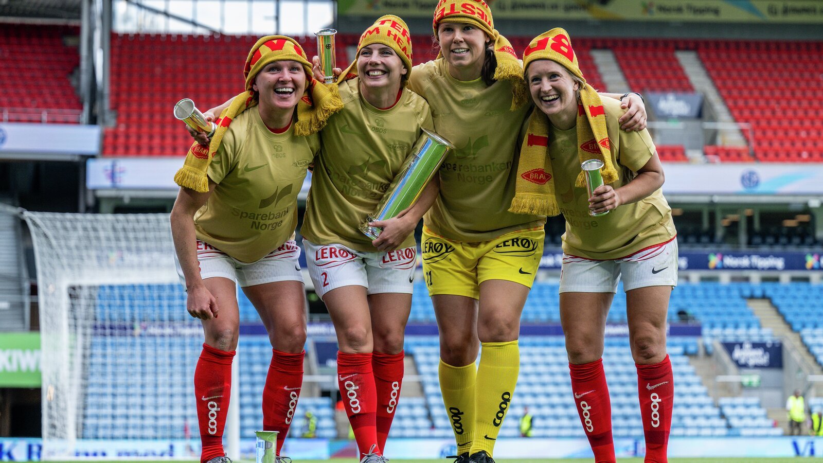 Fem kvinner i matchende røde og gule sportsuniformer feirer sammen på en fotballbane på stadion.